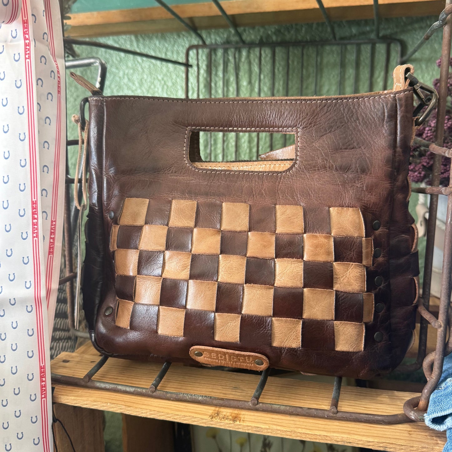 A brown leather handbag with a woven checkerboard pattern panel on the front, displayed on a rustic metal wire shelf.
