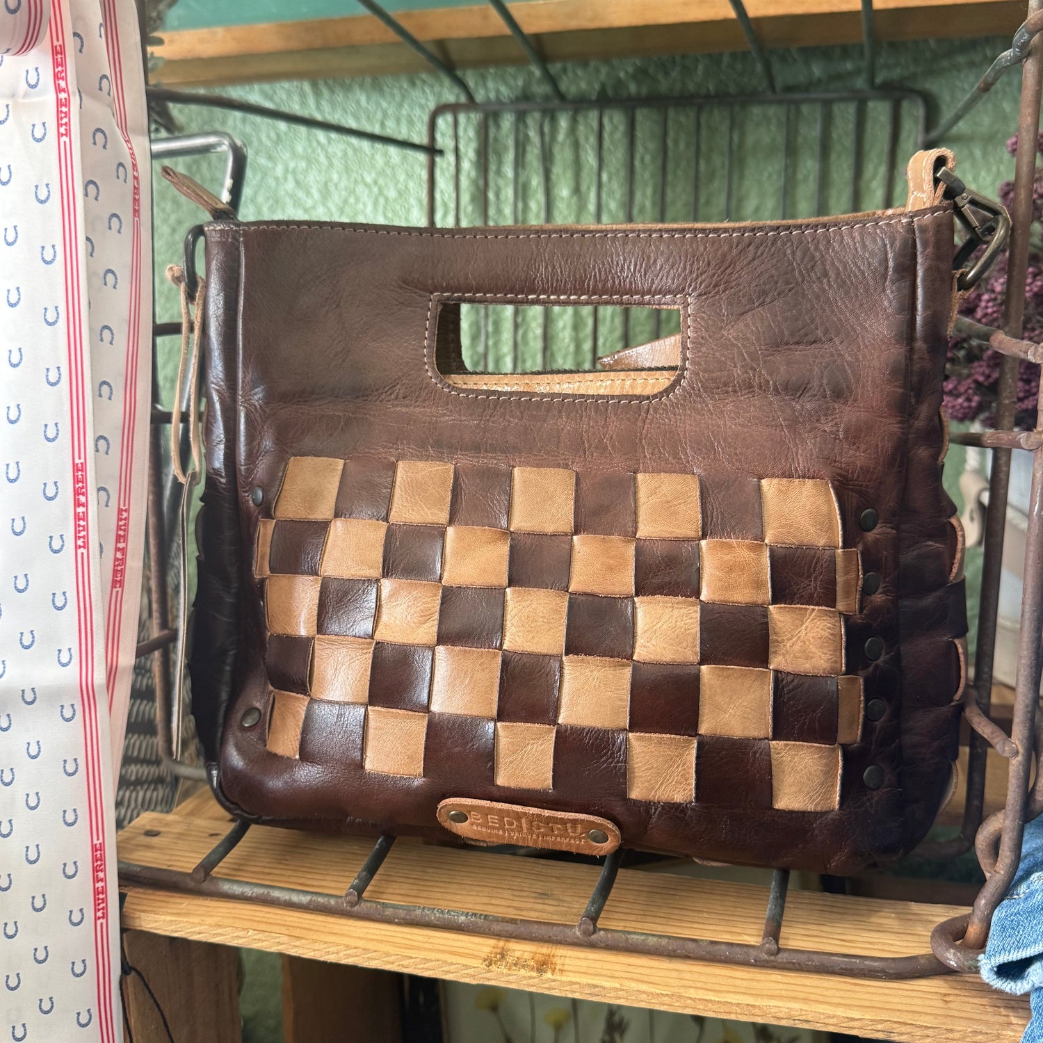 A brown leather handbag with a woven checkerboard pattern panel on the front, displayed on a rustic metal wire shelf.
